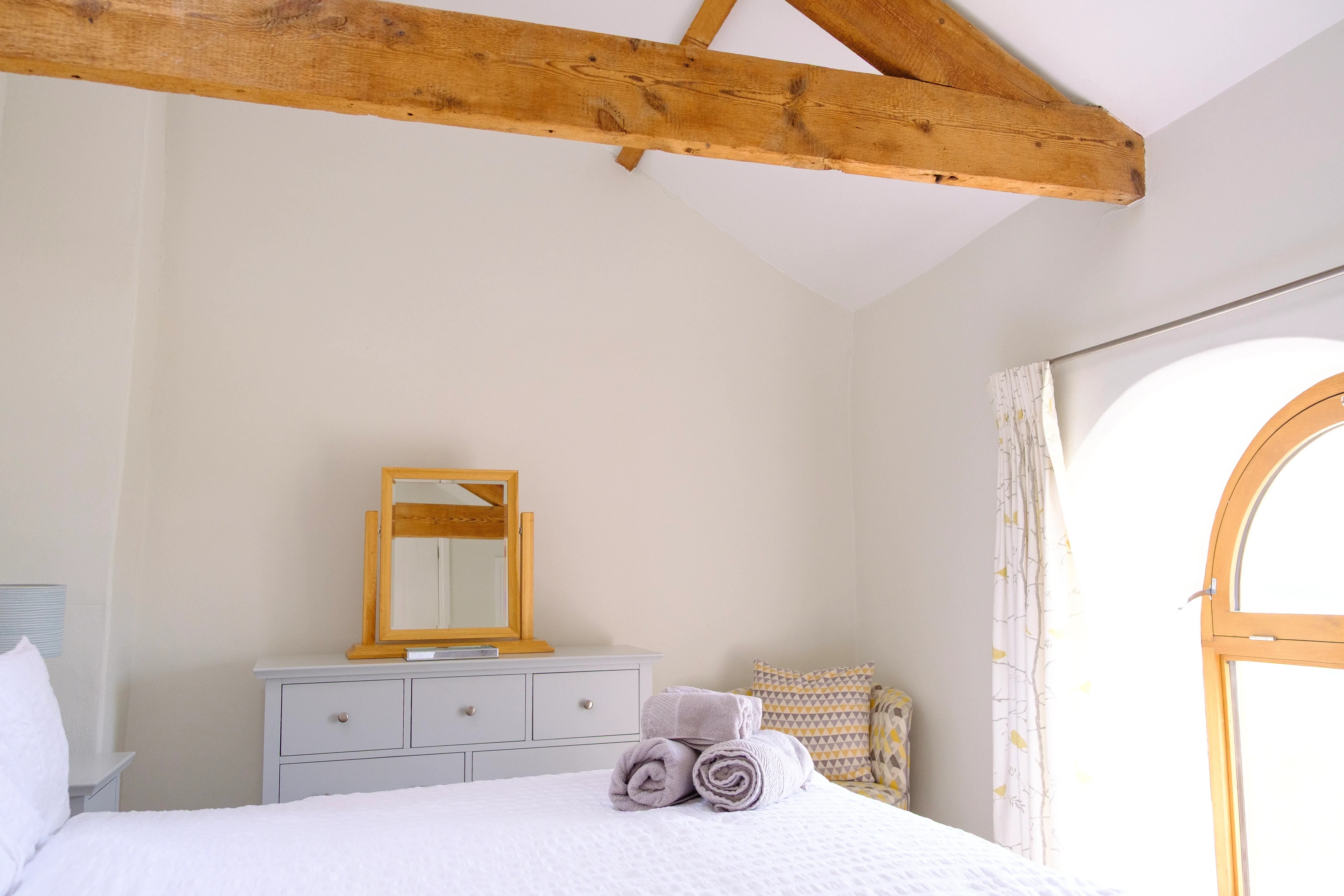 The main bedroom showing airy white walls, exposed beams, a chest of drawers, and a little arm chair in the corner.